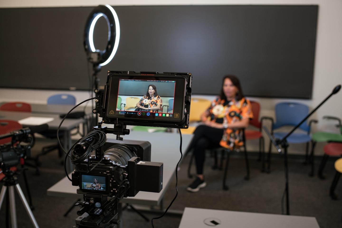 Alumna sitting in chair in classroom being videotaped for interview. 