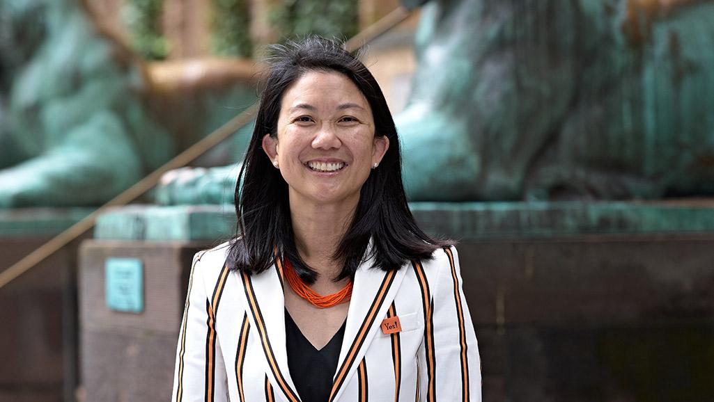 Deb Yu, wearing her class jacket and standing in front of Nassau Hall, with the tigers statues behind her