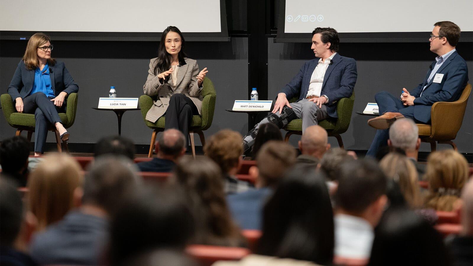 Four people are seated on-stage in front of a crowded audience.